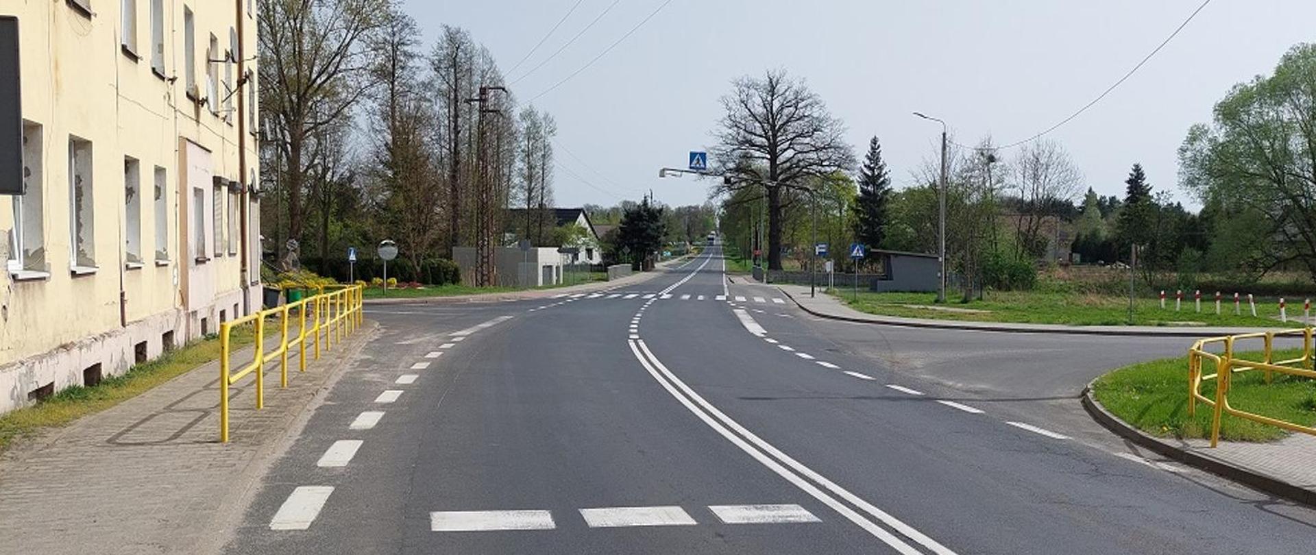 Skrzyżowanie drogi krajowej nr 12 w Lipinkach Łużyckich – dwupasmowa jezdnia z oznakowaniem poziomym, przejściem dla pieszych i wysepką, chodniki z żółtymi barierkami oraz zabudowa po lewej stronie i tereny zielone po prawej.