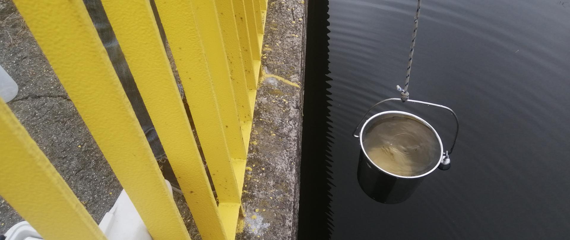 Wiadro z wodą na tle rzeki, obok barierka mostu i sprzęt pomiarowy
