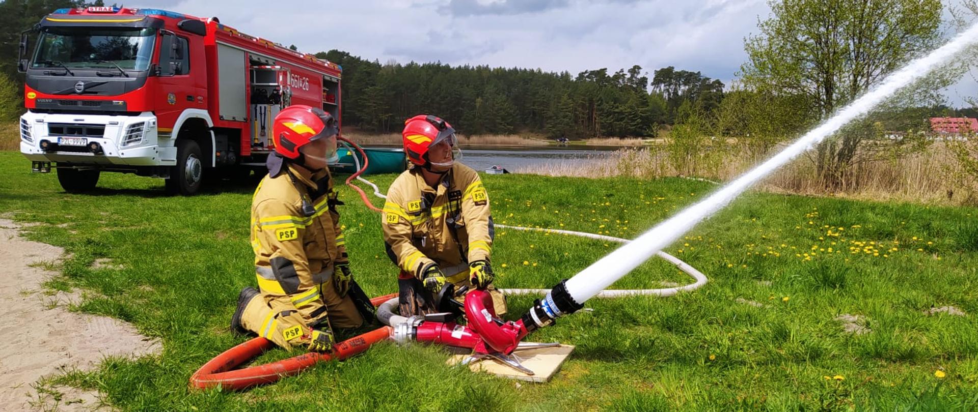 Na zdjęciu strażacy na tle zielonej trawy operują wodą z działka.