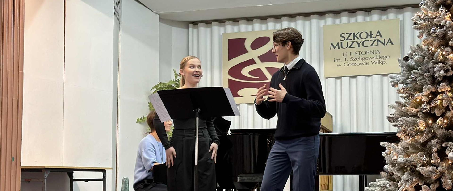 Dziewczyna i chłopak śpiewają na scenie. W tle siedzi pianistka. Z prawej strony przystrojona choinka