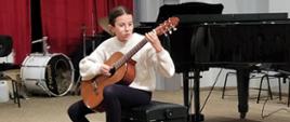 Dziewczynka siedzi na krześle na środku estrady auli PSM i gra na gitarze. Za nią stoi fortepian i zestaw perkusyjny.