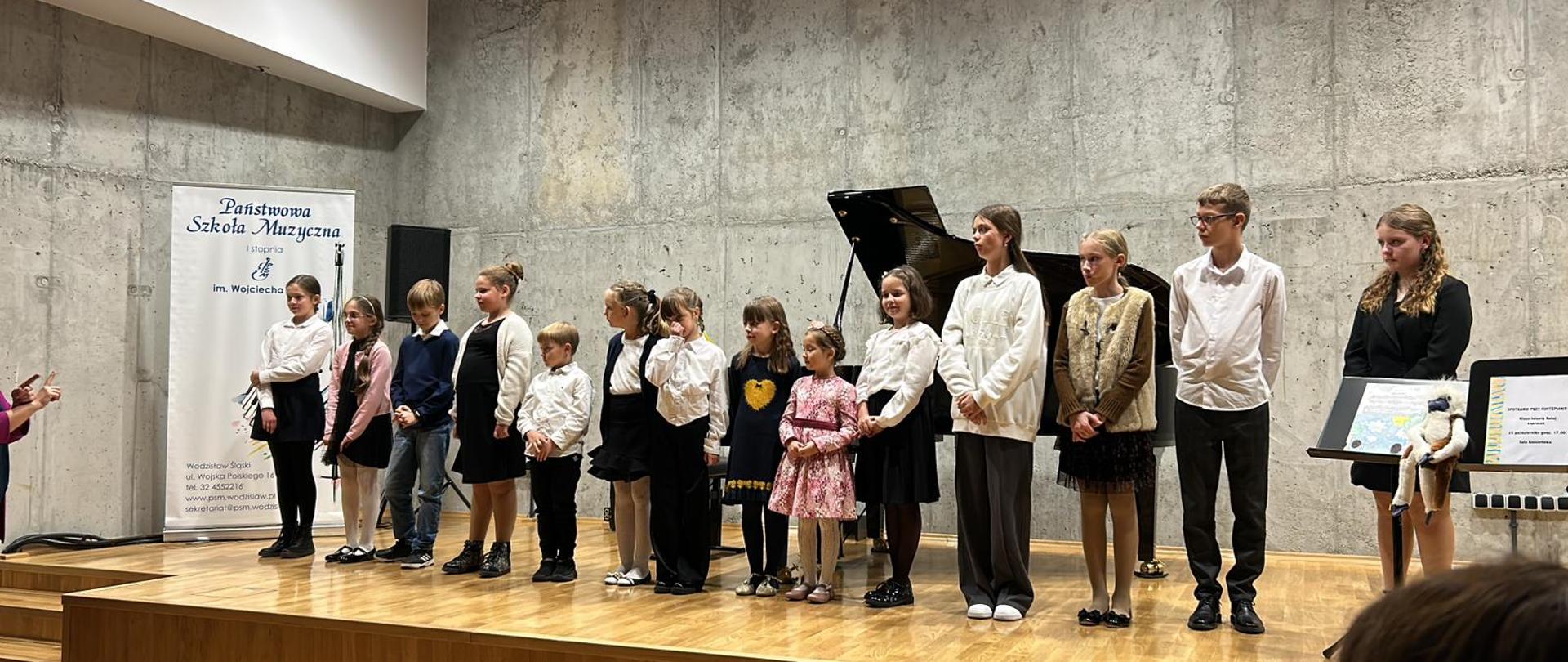 Grupa uczniów stoi na scenie sali koncertowej, fortepian w tle