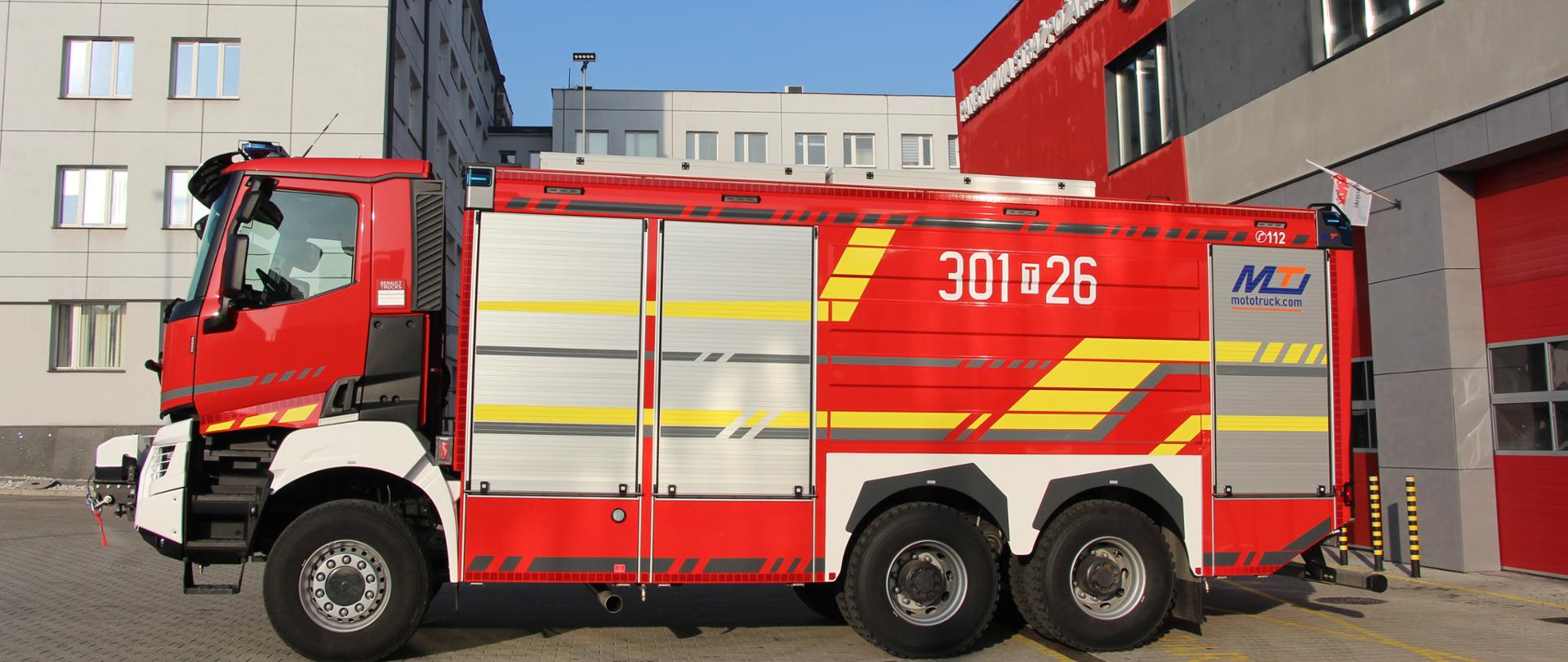 Zdjęcie przedstawia nowy ciężki samochód gaśniczy stojący przed garażem Jednostki Ratowniczo-Gaśniczej Numer 1 w Kielcach. Za nim budynki Komendy Wojewódzkiej i Komendy Miejskiej Państwowej Straży Pożarnej w Kielcach.
