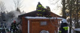 Na zdjęciu widoczne działania strażaków psp i osp przy domku letniskowym objętym pożarem. widoczny śnieg, strażacy pracujący na dachu obiektu. 