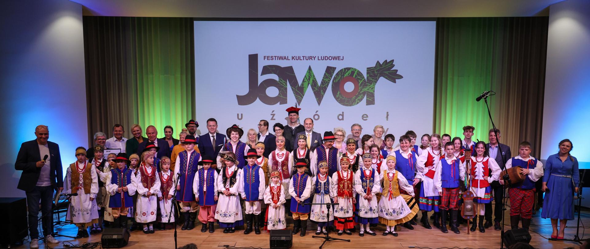 Zbiorowe zdjęcie uczestników festiwalu kultury ludowej "Jawor- u źródeł kultury". Uczestnicy w strojach ludowych. W tle na białym polu nazwa uroczystości.