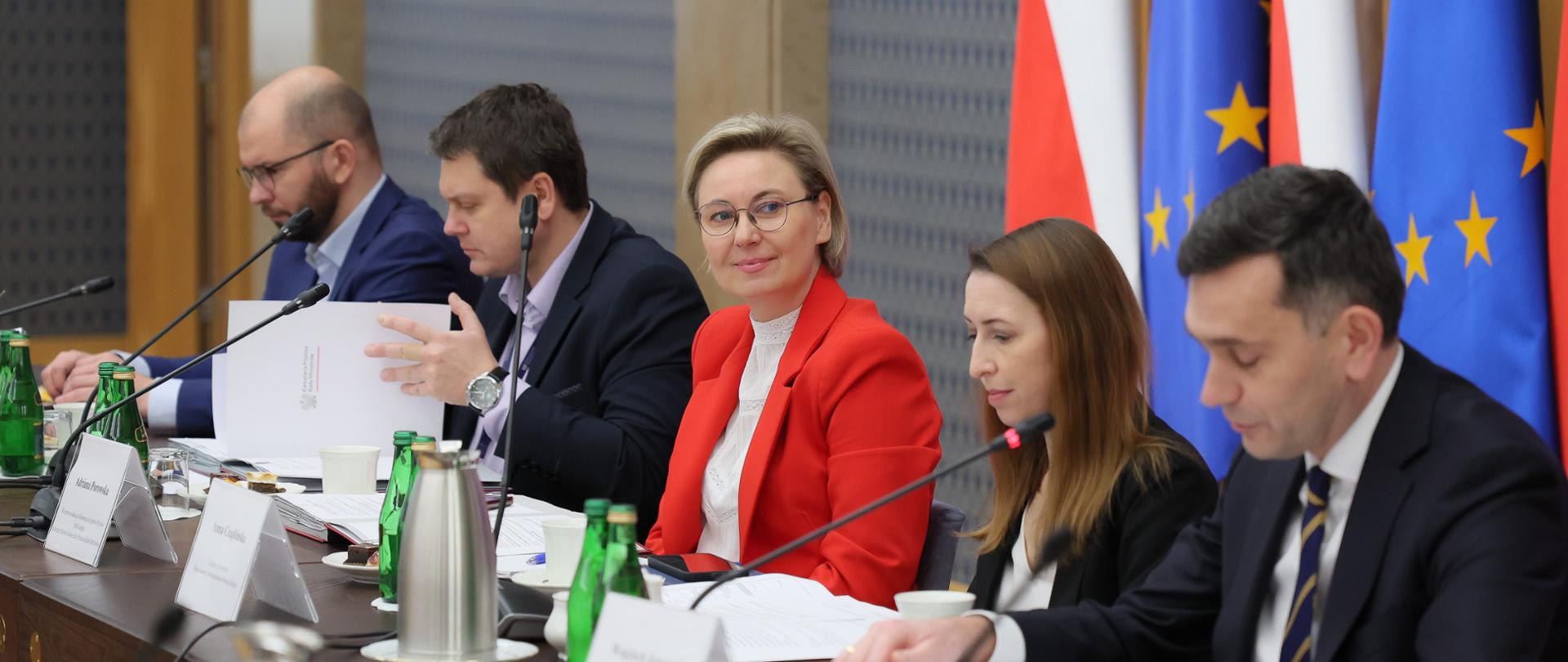 Na zdjęciu uczestnicy przy stole konferencyjnym. Od lewej: Pan Michał Braun, Dyrektor Narodowego Instytutu Wolności – Centrum Rozwoju Społeczeństwa Obywatelskiego, Pan Łukasz Jachimowicz, Dyrektor Departamentu Społeczeństwa Obywatelskiego w KPRM, Pani Adriana Porowska, Sekretarz Stanu w KPRM, Wiceprzewodnicząca Komitetu do Spraw Pożytku Publicznego oraz zastępcy dyrektora Departamentu Społeczeństwa Obywatelskiego w KPRM Pani Anna Czaplińska i Pan Wojciech Kuraszyk. W tle flagi Polski i Unii Europejskiej.