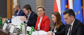 Na zdjęciu uczestnicy przy stole konferencyjnym. Od lewej: Pan Michał Braun, Dyrektor Narodowego Instytutu Wolności – Centrum Rozwoju Społeczeństwa Obywatelskiego, Pan Łukasz Jachimowicz, Dyrektor Departamentu Społeczeństwa Obywatelskiego w KPRM, Pani Adriana Porowska, Sekretarz Stanu w KPRM, Wiceprzewodnicząca Komitetu do Spraw Pożytku Publicznego oraz zastępcy dyrektora Departamentu Społeczeństwa Obywatelskiego w KPRM Pani Anna Czaplińska i Pan Wojciech Kuraszyk. W tle flagi Polski i Unii Europejskiej.