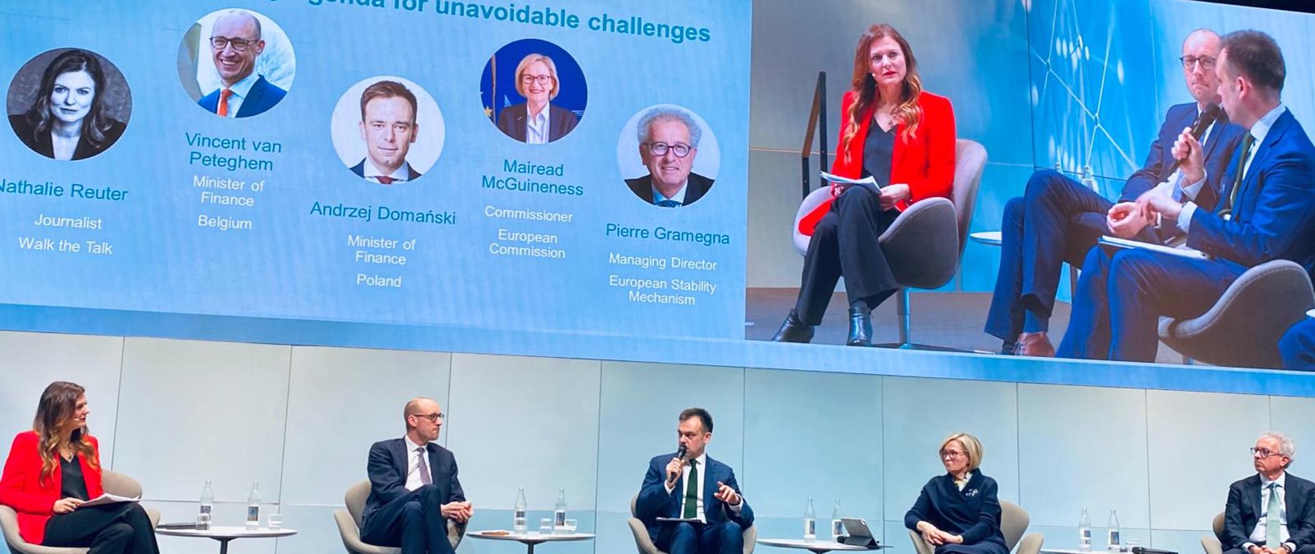 Minister finansów Andrzej Domański w otoczeniu panelistów, mówi do mikrofonu, podczas Forum Grupy Europejskiego Banku Inwestycyjnego w Luksemburgu, w tle telebim z uczestnikami