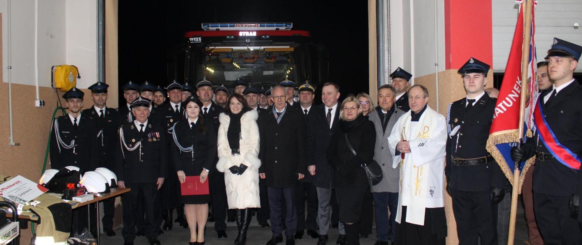 Zdjęcie grupowe uczestników uroczystości w pomieszczeniu garażu na tle otwartej bramy, w której widoczny jest przód samochodu strażackiego. Na pierwszym planie po lewej stronie stoi stolik z ułożonym asortymentem wyposażenia osobistego strażaków w postaci butów, hełmów, i ubrania bojowego. Po prawej stronie stoi poczet sztandarowy jednostki OSP. 