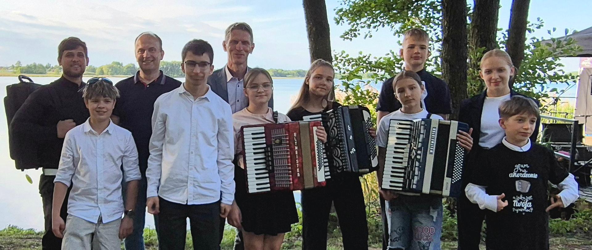 grupa dzieci i dorosłych stojąca na trawie, niektórzy trzymają instrumenty muzyczne, w tle drzewa i jezioro
