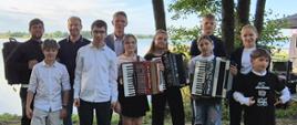 grupa dzieci i dorosłych stojąca na trawie, niektórzy trzymają instrumenty muzyczne, w tle drzewa i jezioro