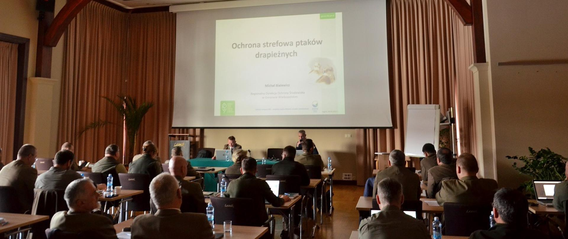 Ogólny widok sali, w której na krzesłach, odwróceni tyłem siedzi kilkadziesiąt osób. W głębi sali za stołem przodem do pozostałych siedzi dwóch mężczyzn. Na ścianie za ich plecami wisi ekran, na którym znajduje się slajd prezentacji. Na nim napis: Ochrona strefowa ptaków drapieżnych.