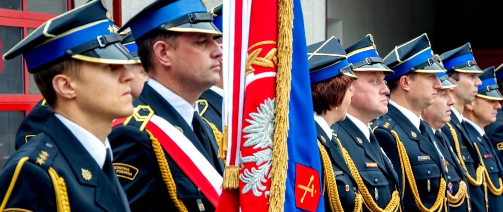 Na zdjęciu widać poczet sztandarowy oraz funkcjonariuszy Państwowej Straży Pożarnej w mundurach galowych ustawionych w rzędzie. 