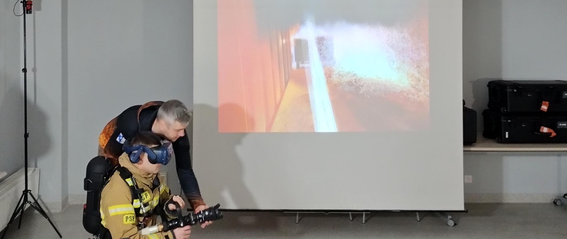 klęczący strażak w googlach i z prądownicą w reku, przy nim pochyla się mężczyzna, za nimi duży ekraz z fotografią pożaru