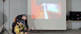 klęczący strażak w googlach i z prądownicą w reku, przy nim pochyla się mężczyzna, za nimi duży ekraz z fotografią pożaru