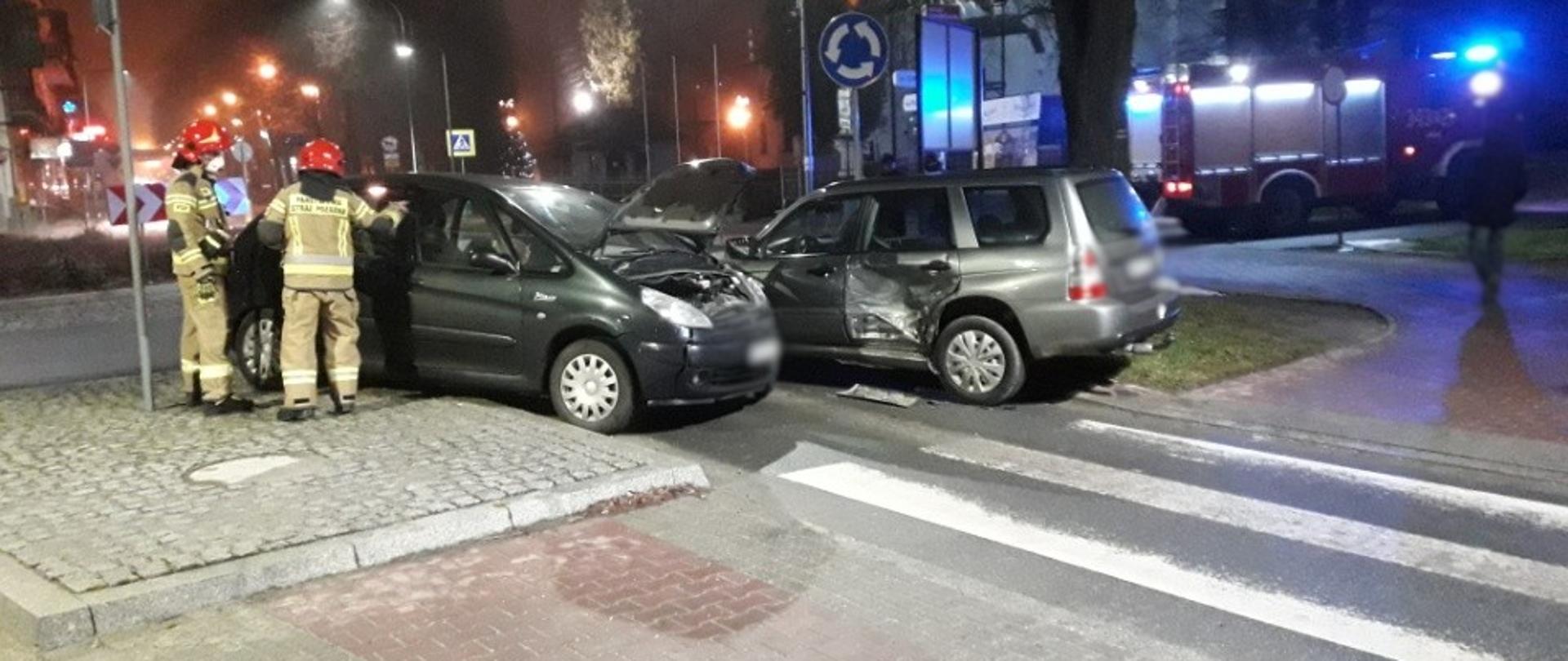 Dwa rozbite samochody stoją na drodze przed wjazdem na rondo. Obok samochodu stoi kilku strażaków i prowadzi swoje działania.