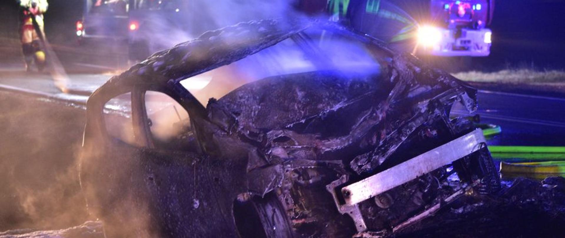 Na zdjęciu widać spalony samochód osobowy. Z auta unosi się para wodna, jest pora nocna. W tle widać samochód pożarniczy oraz strażaków zwijających sprzęt po akcji