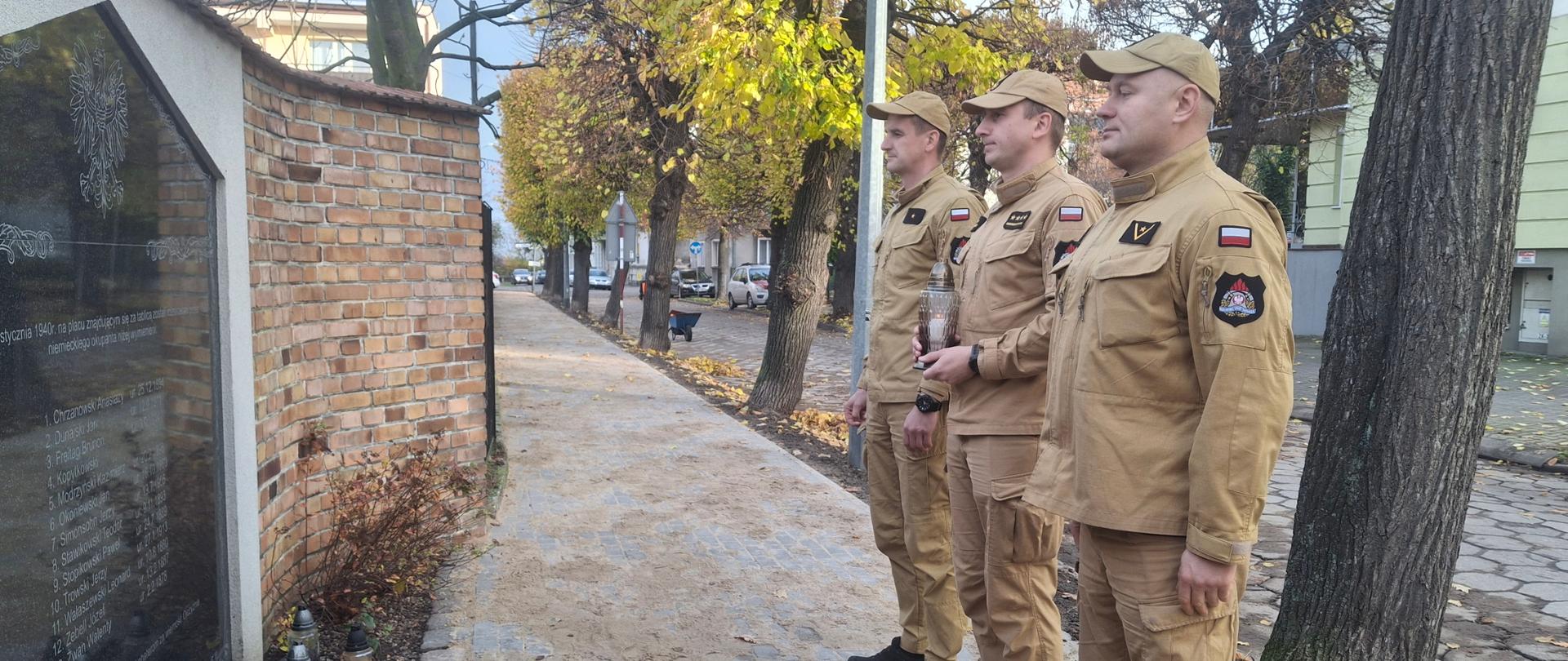 Upamiętnienie ofiar II Wojny Światowej - trzech strażaków ze zniczem przed pomnikiem,