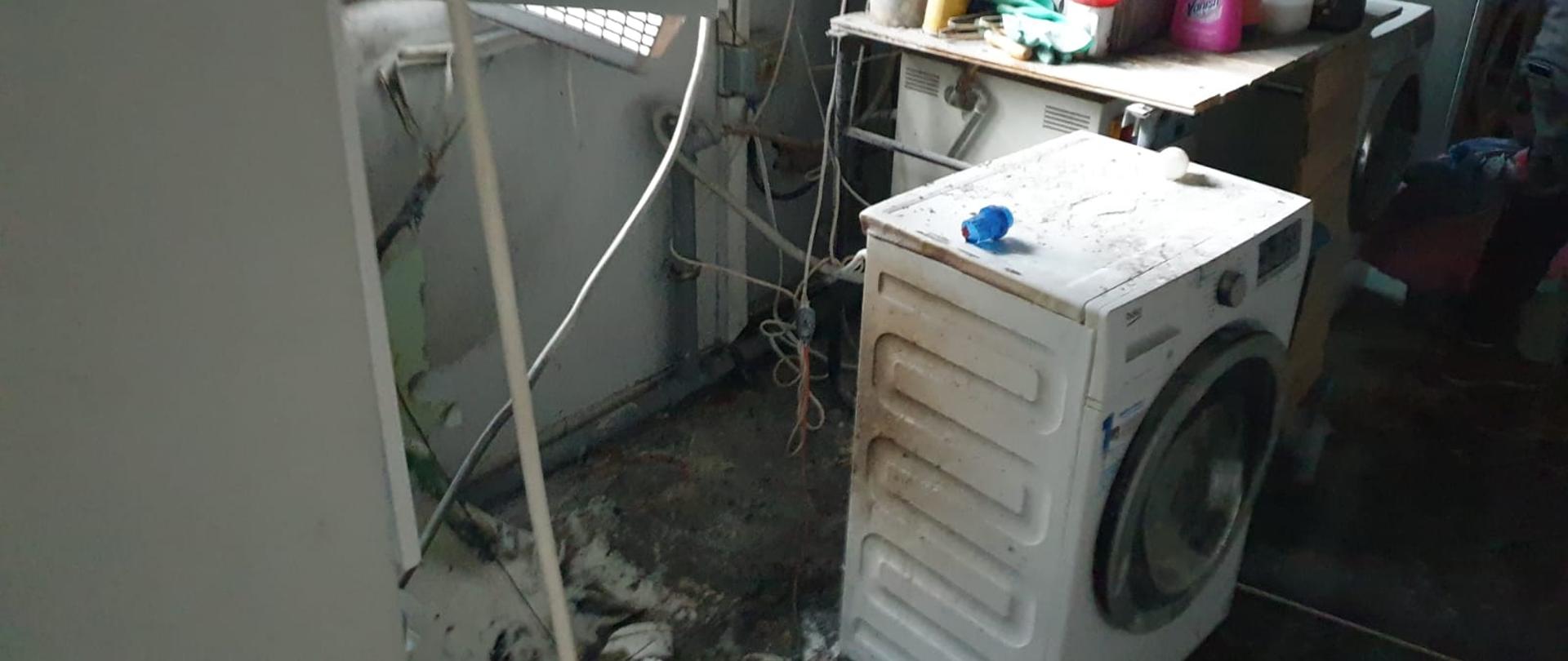 Na zdjęciu widać miejsce pożaru w pralni. Uszkodzone, stopione instalacje elektryczne oraz pralka. Okopcone ściany. 