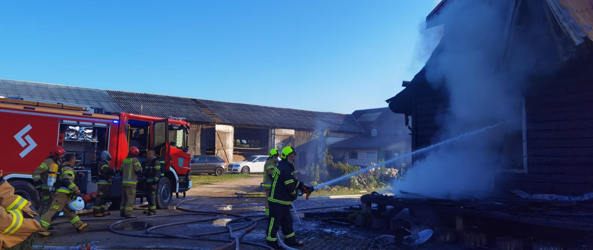 Strażak leje wodę z węża strażackiego przez okno dymiącego budynku. W tle samochód strażacki oraz stojący przy nim strażacy 