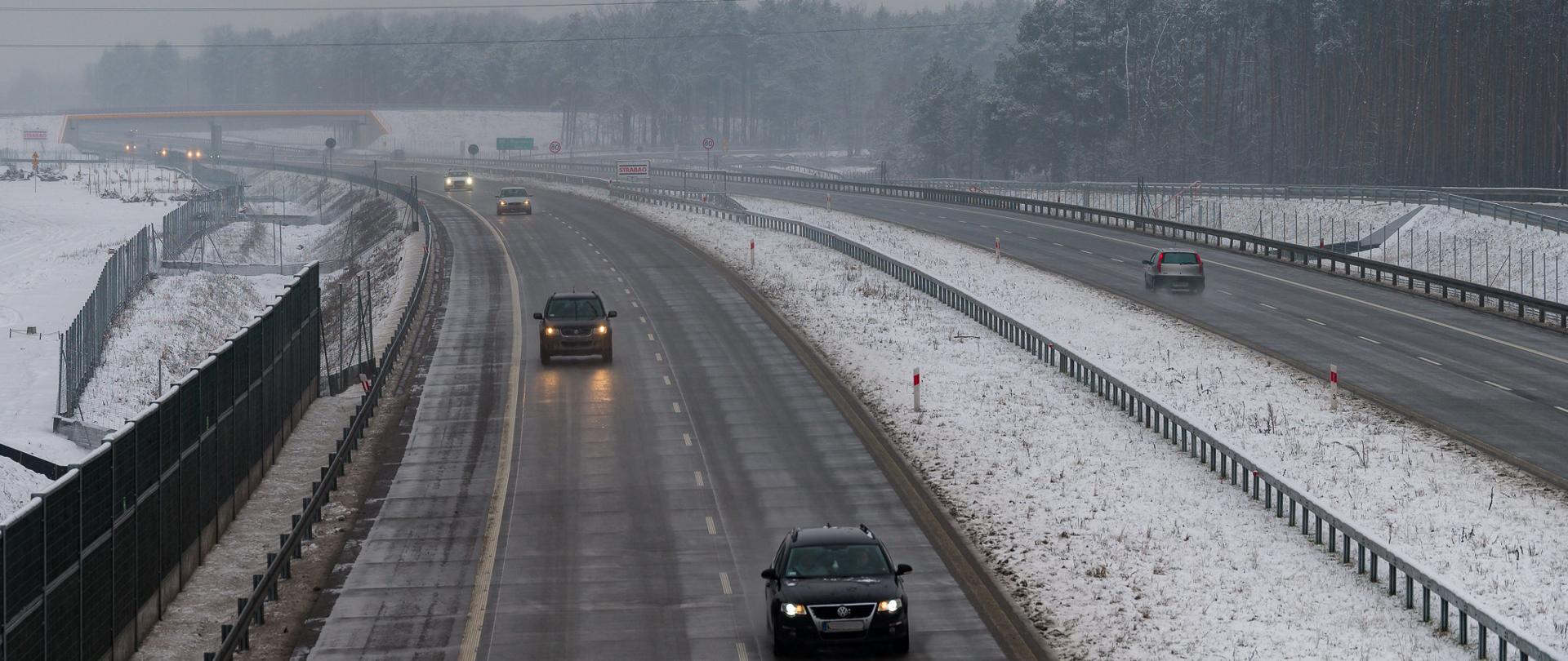 Zimowy widok na dwupasmową drogę ekspresową otoczoną lasem, z kilkoma samochodami jadącymi w obu kierunkach. Jezdnia jest mokra, a pobocza i otoczenie pokryte cienką warstwą śniegu. W tle widoczne są ekrany akustyczne, znaki drogowe oraz las sosnowy pod pochmurnym, szarym niebem.