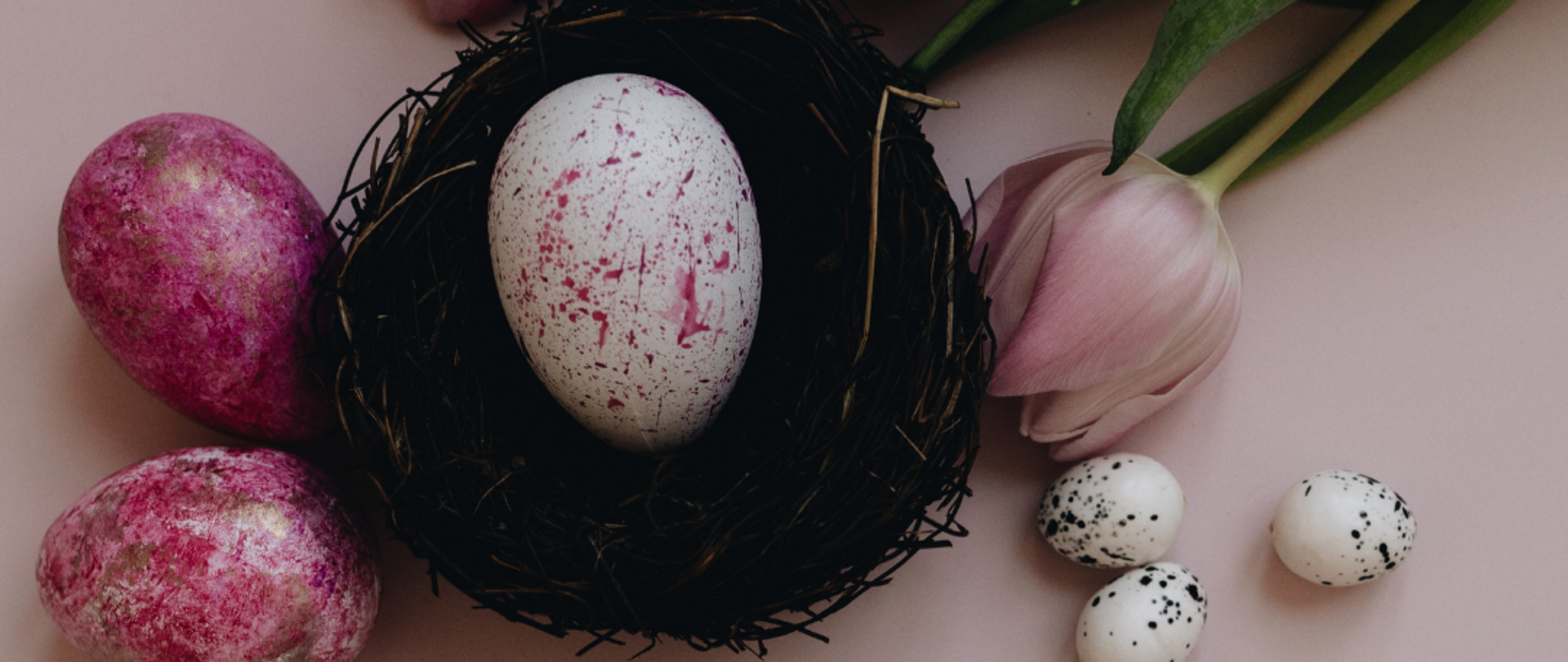 Na różowym tle ułożone w kompozycje różowe tulipany oraz biało-różowe pisanki . Na środku napis Wesołych Świąt Wielkanocnych.