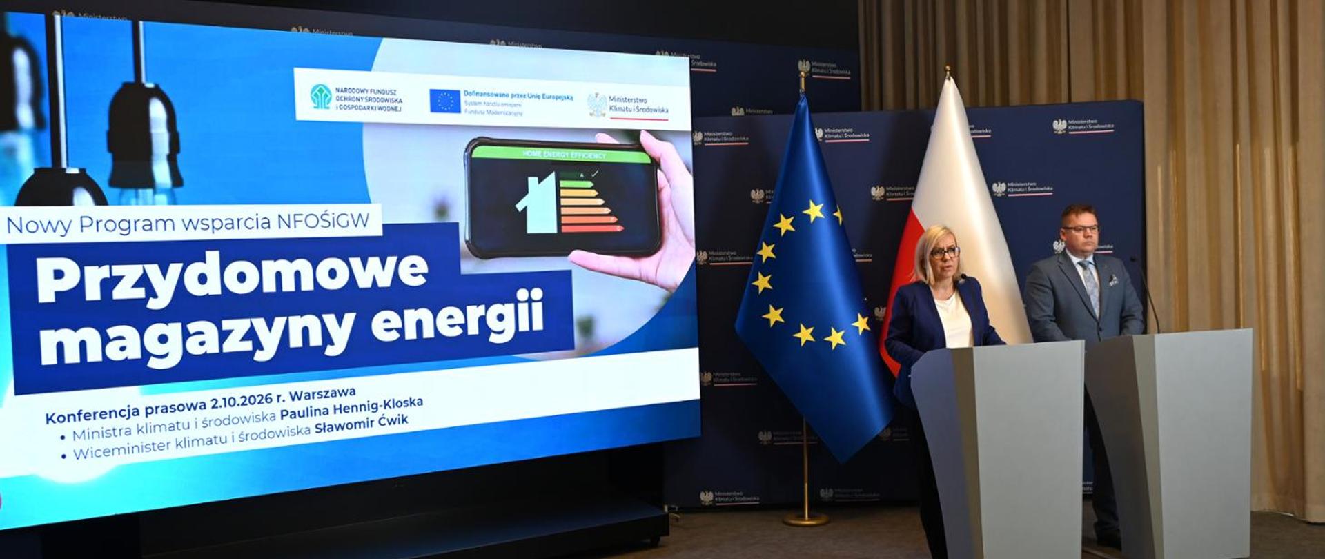 Sala konferencyjna z dużym ekranem prezentacyjnym zawierającym napis „Przydomowe magazyny energii” oraz grafikę przedstawiającą klasę efektywności energetycznej budynku. Obok ekranu znajdują się mównice, przy których stoją uczestnicy wydarzenia. W tle widoczne są flagi Polski i Unii Europejskiej oraz ścianka Ministerstwa Klimatu i Środowiska.