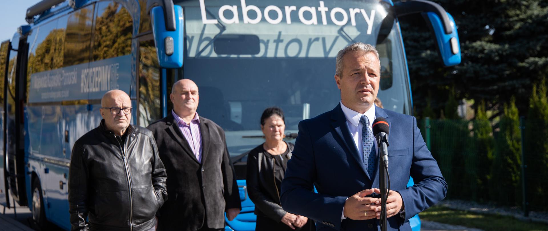 Briefing prasowy rozpoczynający trasę szczepionkobusa w powiecie inowrocławskim - gmina Janikowo. Na pierwszym planie stoi mężczyzna przy mikrofonie. 