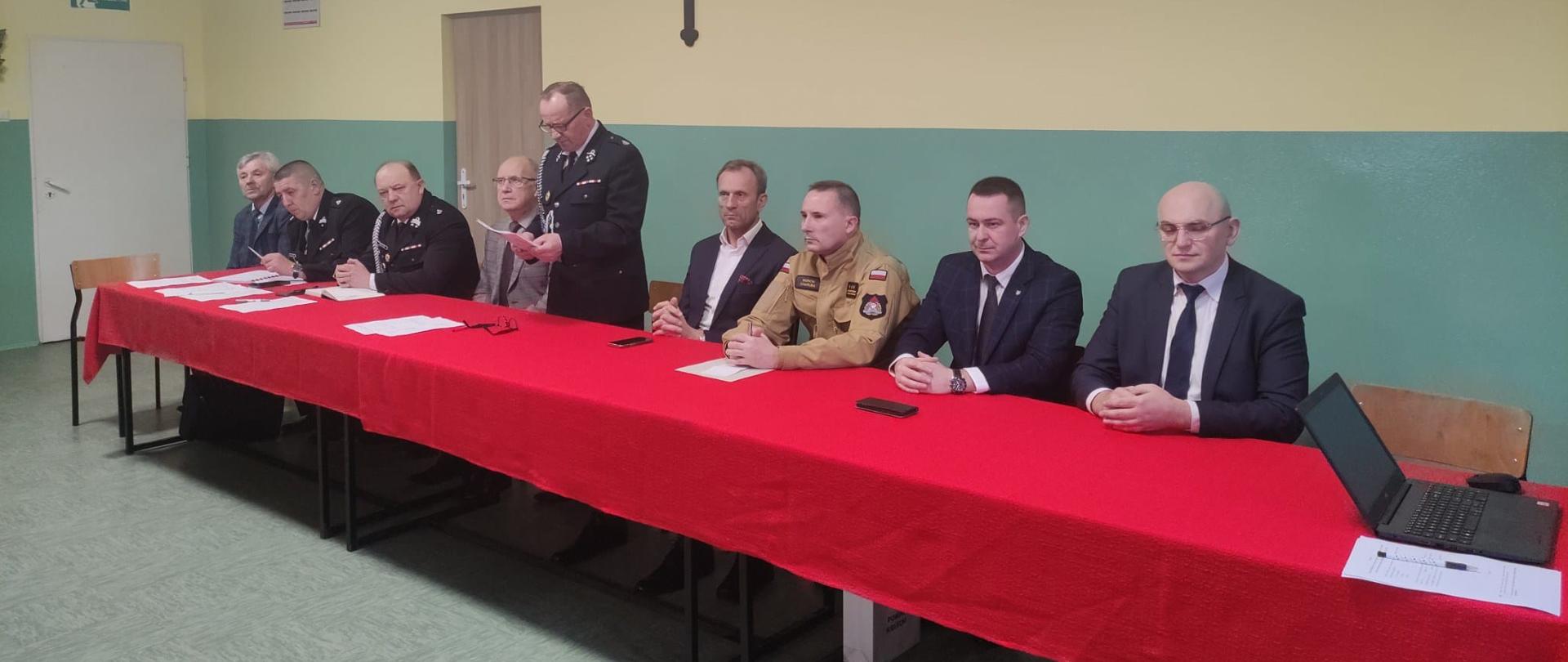 Zdjęcie przedstawia organizatorów i gości siedzący przy stole podczas spotkania sprawozdawczego Ochotniczej Straży Pożarnej w Rykoszynie.