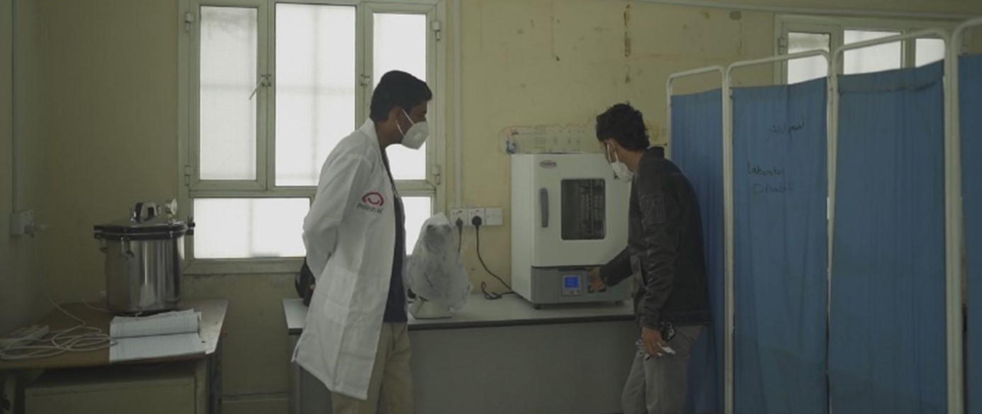 Dwóch mężczyzn stoi przy urządzeniu do prowadzenia badań. Obok ustawiono niebieski parawan.