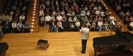 Widok na zapełnioną widownię, z przodu estrada, na której tyłem (a przodem do widowni) stoi mężczyzna i mówi do mikrofonu. Po prawej stronie stoi fortepian.