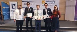 Pod ścianą na której wisi baner z napisem Economics Olympiad stoi czterech mężczyzn i kobieta, trzech w środku trzyma dyplomy.