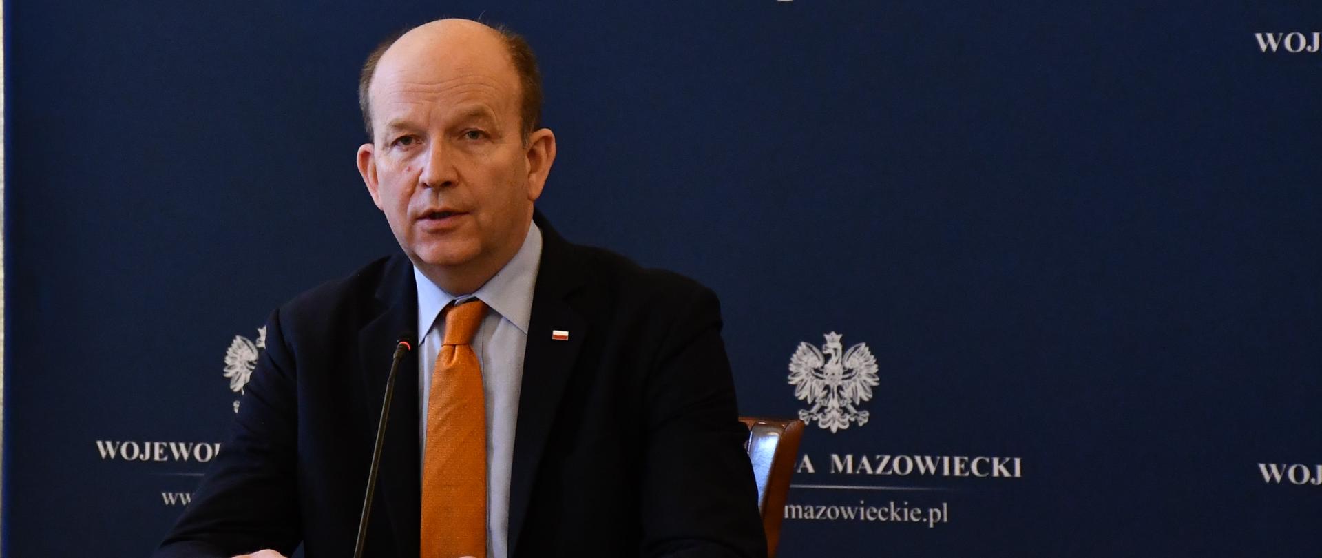 Elegancko ubrany mężczyzna przemawia podczas wideokonferencji. W tle znajduje się granatowe tło z białymi napisami i orłem