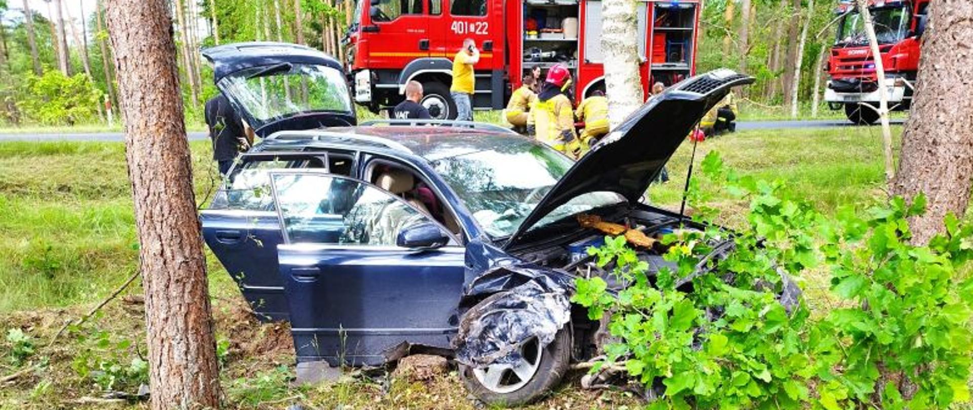 Zdjęcie wykonane w letni dzień. Centralny punkt stanowi rozbite auto które przodem wjechało w krzaki. Auto ma ciemny kolor. W tle wzdłuż drogi stoją pojazdy służba ratowniczych.