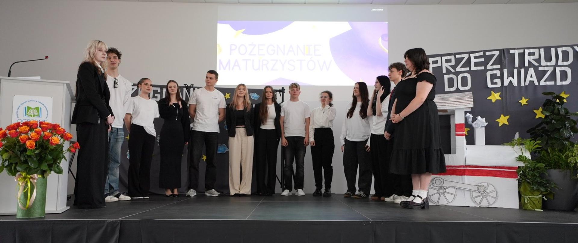  Grupa osób stoi na scenie podczas uroczystości; w tle projekcja „Pożegnanie Maturzystów” i dekoracja „Przez trudy do gwiazd”.