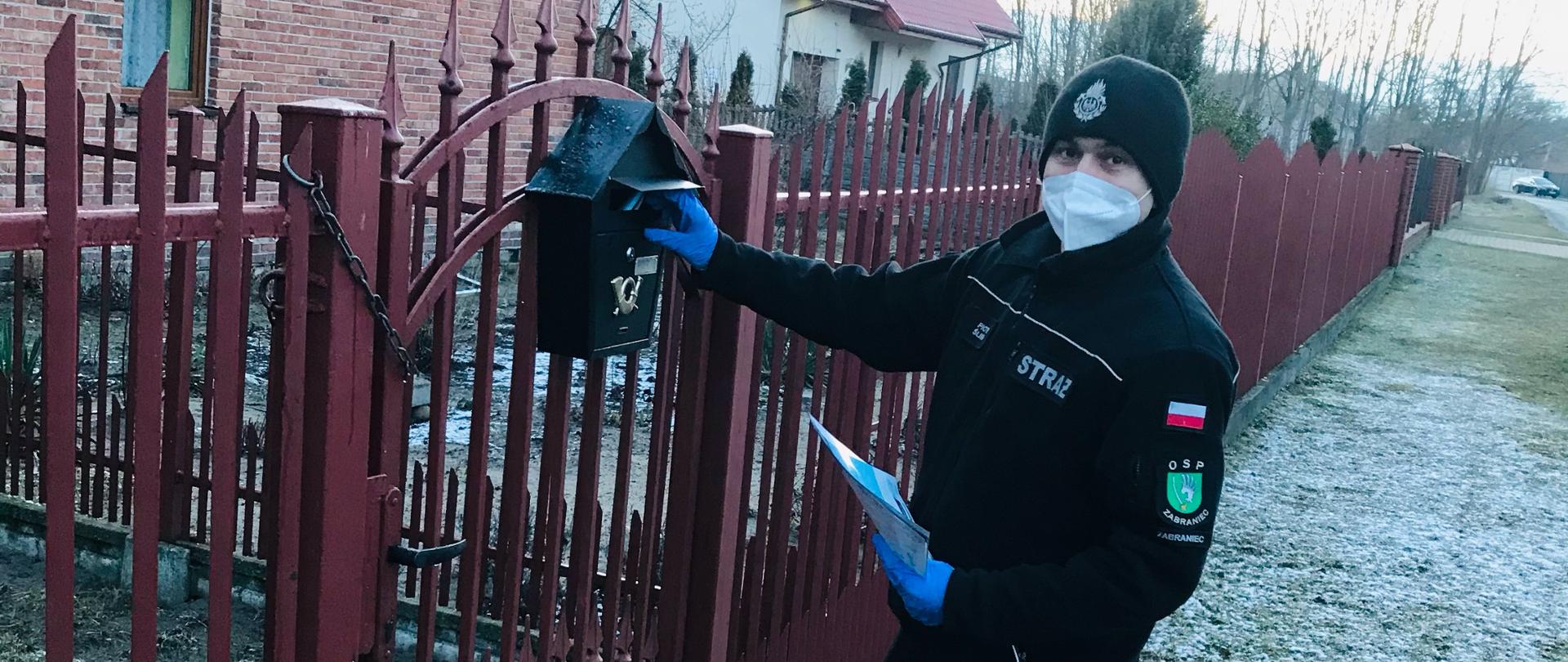 Na zdjęciu druh Ochotniczej Straży Pożarnej ubrany w umundurowanie bojowe wkładający ulotkę do skrzynki na listy zawieszonej na ogrodzeniu posesji.