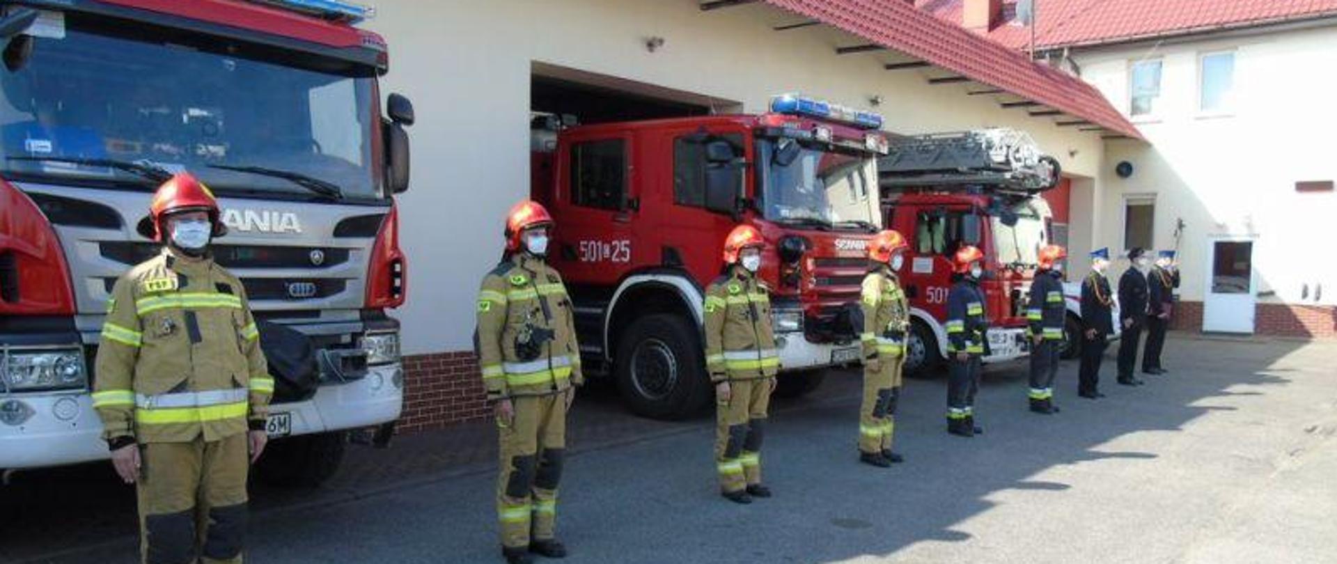 Zdjęcie strażaków w umundurowaniu i maseczkach ochronnych na tle samochodów pożarniczych.