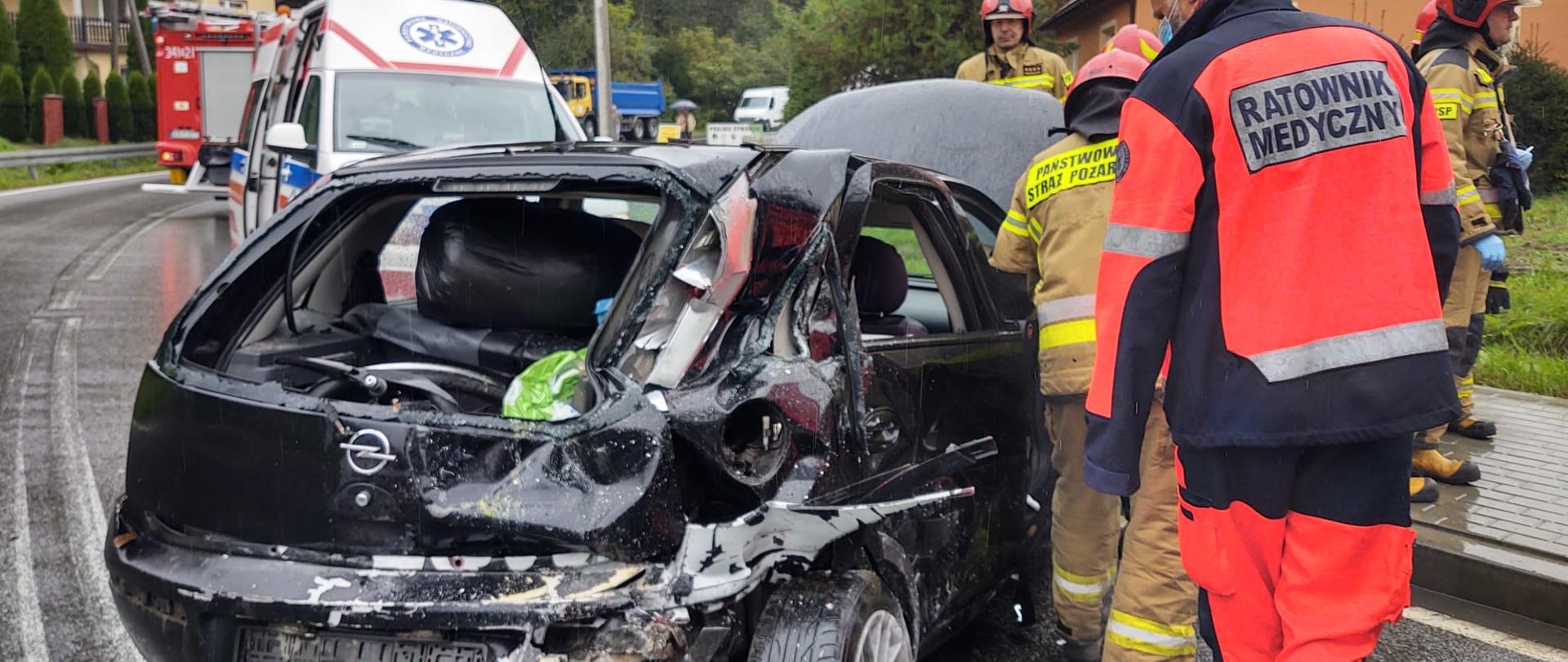 Zniszczony samochód osobowy. W tle karetka. Ratownik medyczny i strażacy prowadzący działania