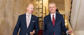 Zdjęcie przedstawia idących po schodach ministrów: Tomasza Siemoniaka (po prawej) oraz Igora Taro (po lewej). Na schodach czerwony dywan. W tle wnętrza korytarzy KPRM oraz inne osoby idące za ministrami.
