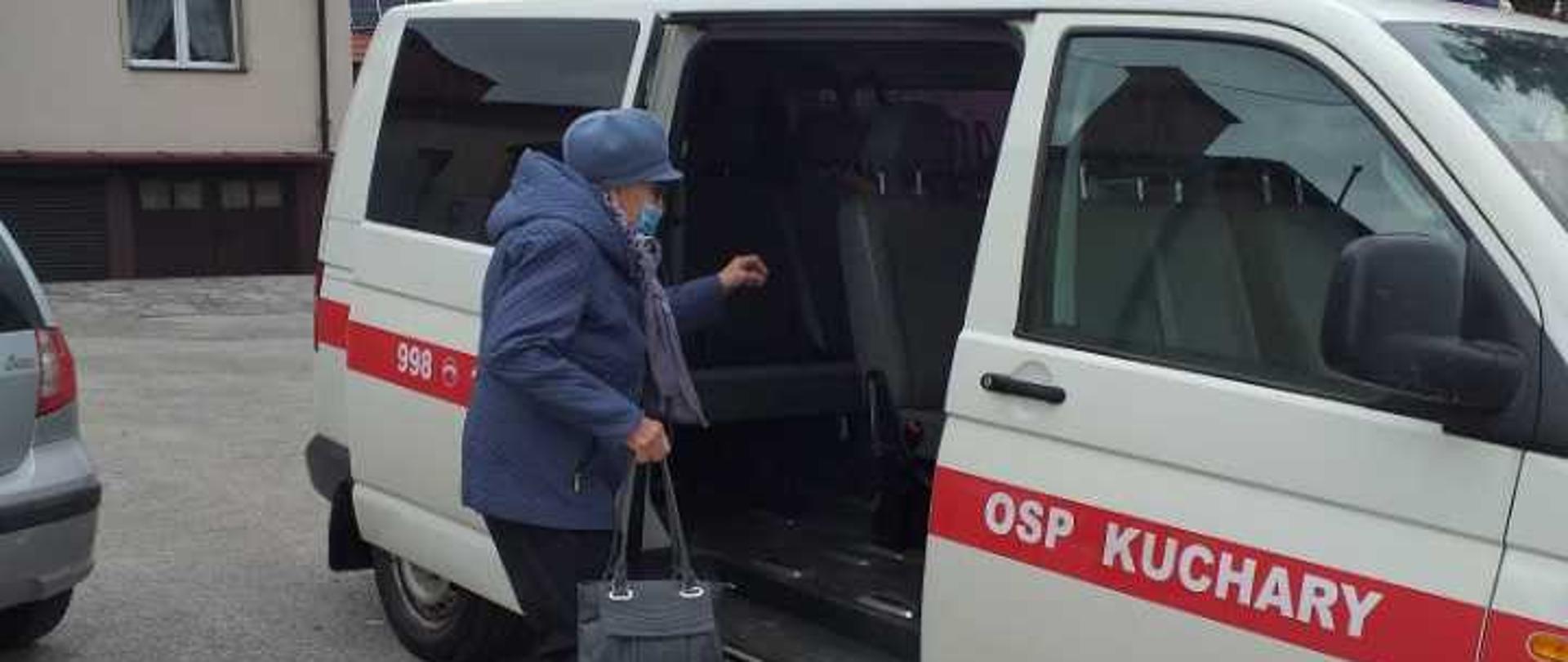 Zdjęcie przedstawiające wchodzącą kobietę do samochodu strażackiego. Samochód koloru białego z czerwonym pasem i napisem OSP Kuchary. W tle budynek.