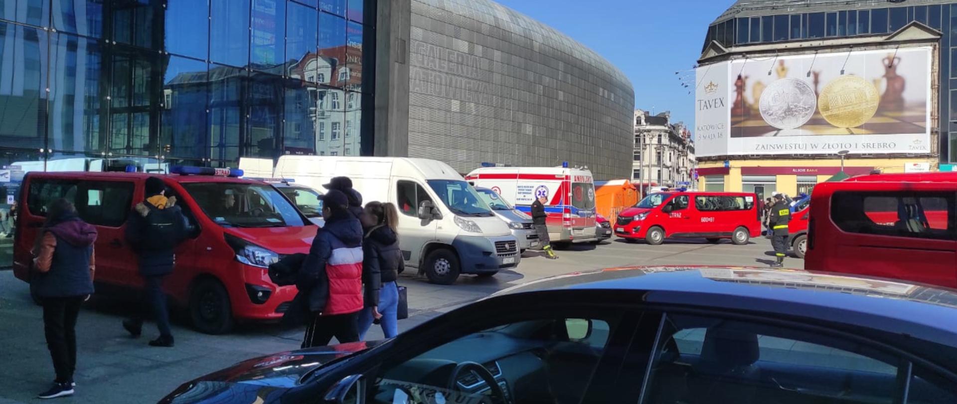Zdjęcie przedstawia kilkanaście samochodów typu bus stojących obok galerii handlowej