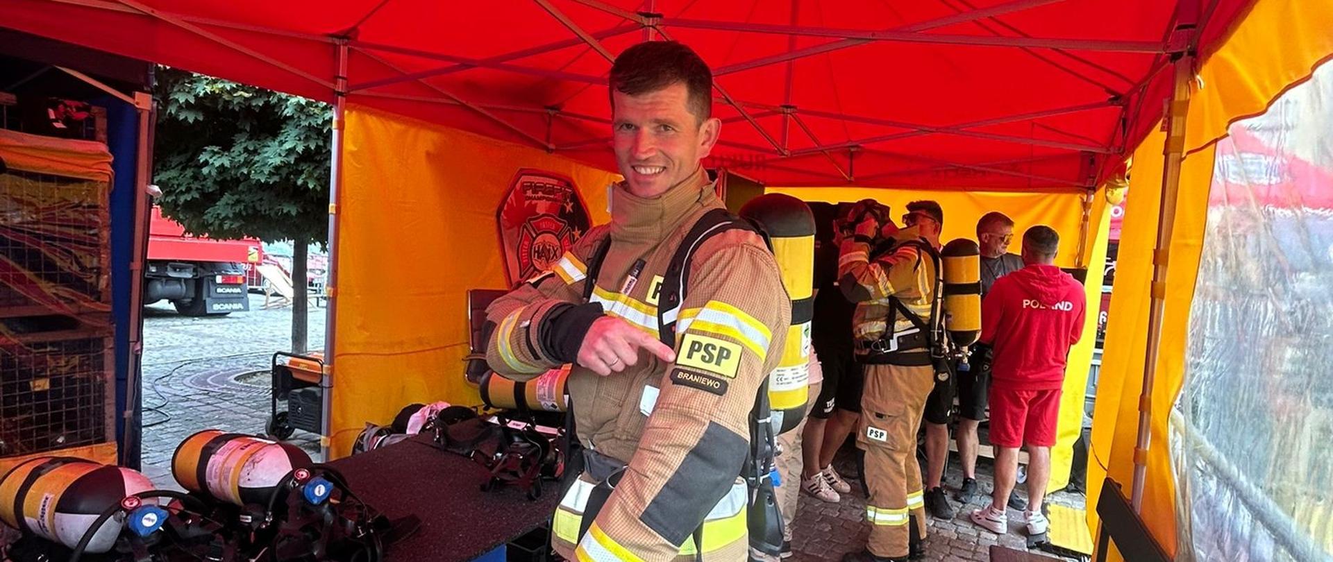 Strażak w mundurze bojowym od czerwonym namiotem. Trwają zawody sportowe strażaków.