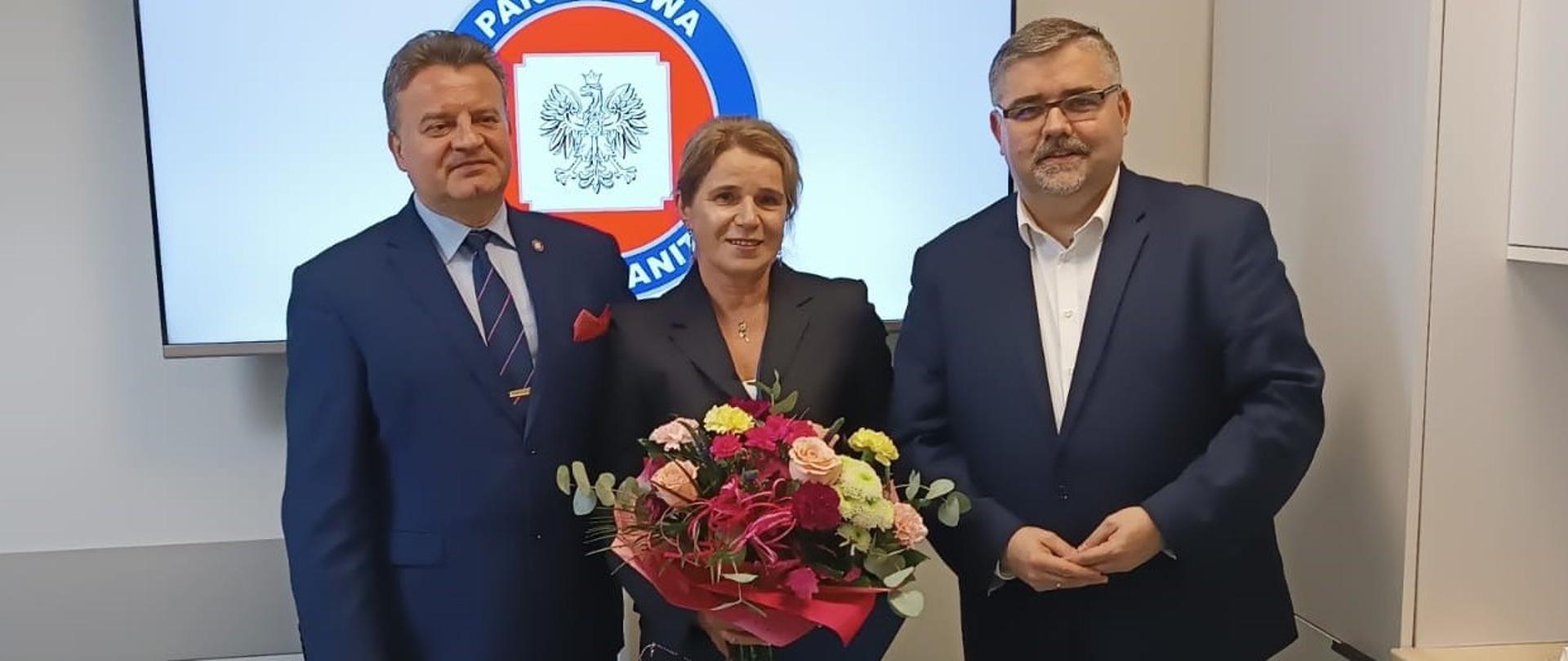 Od lewej stoi mężczyzna w garniturze, obok niego kobieta z bukietem kwiatów w rękach, po prawej stoi drugi meżczyzna