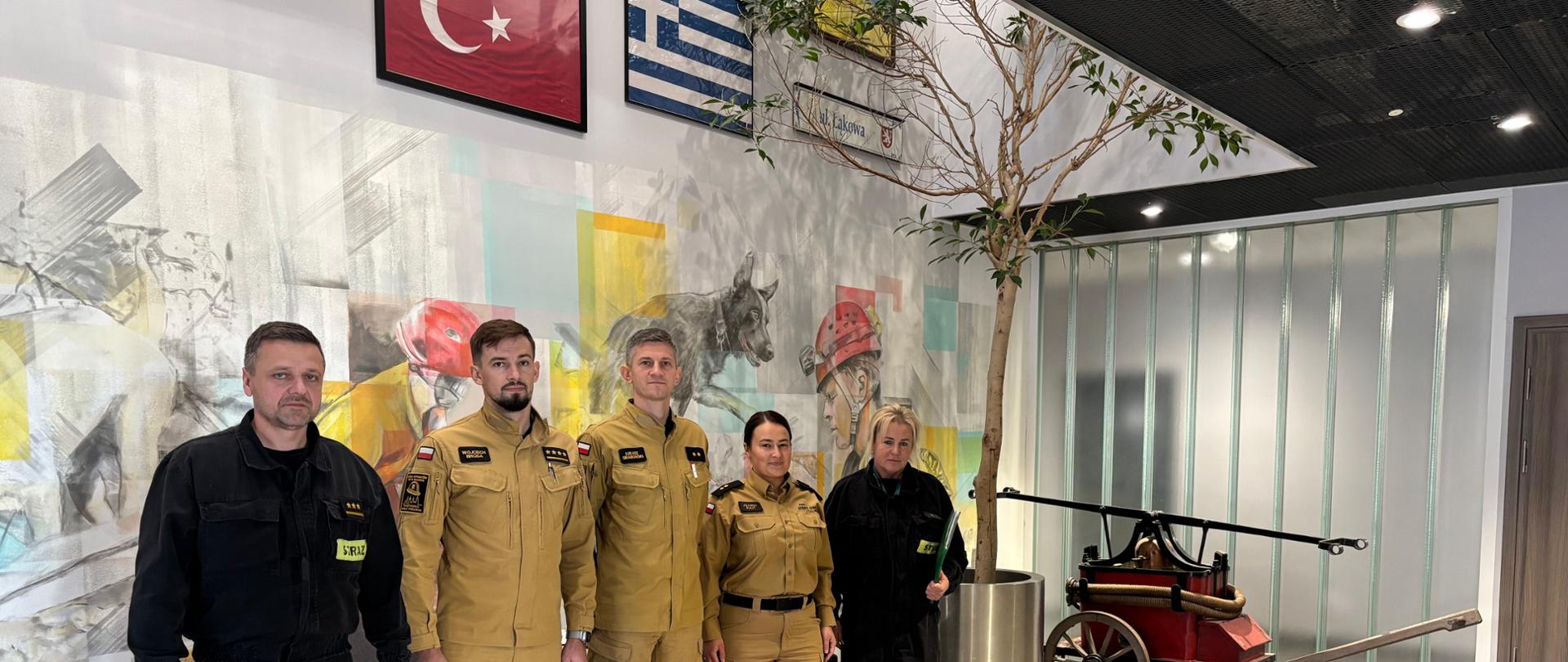 Na zdjęciu przy stołach siedzą 4 osoby-funkcjonariusze PSP. 3 Osoby siedzą plecami do ściany, na której jest zawieszony telewizor. Te osoby to członkowie komisji egzaminacyjnej. Dwóch z nim to mężczyźni przeglądający dokumentację. Trzecia to kobieta patrząca w obiektyw. Przed nimi siedzi mężczyzna w musztardowym mundurze- uczestnik egzaminu. Mężczyzna.