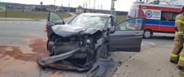 Rozbity szary samochód osobowy z otwartymi drzwiami, ustawiony przodem. Za samochodem w prawej części zdjęcia karetka pogotowia. W oddali osiedle domków jednorodzinnych.
