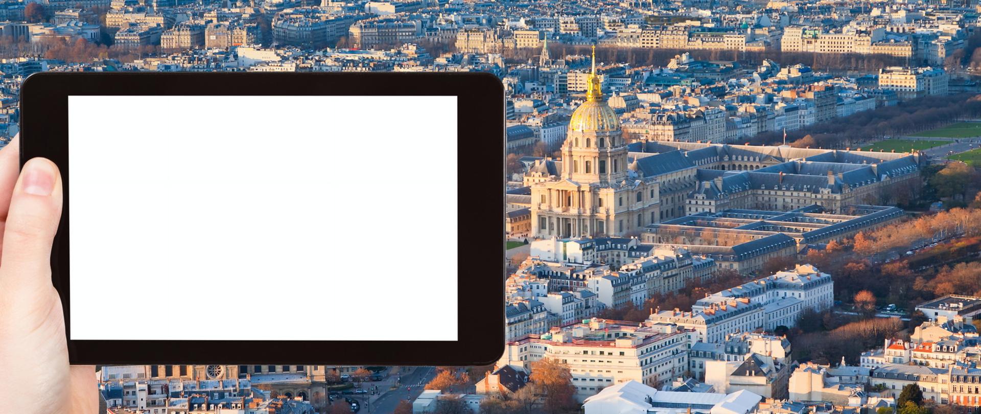Tablet z białym ekranem trzymany w lewej dłoni na tle panoramy miasta