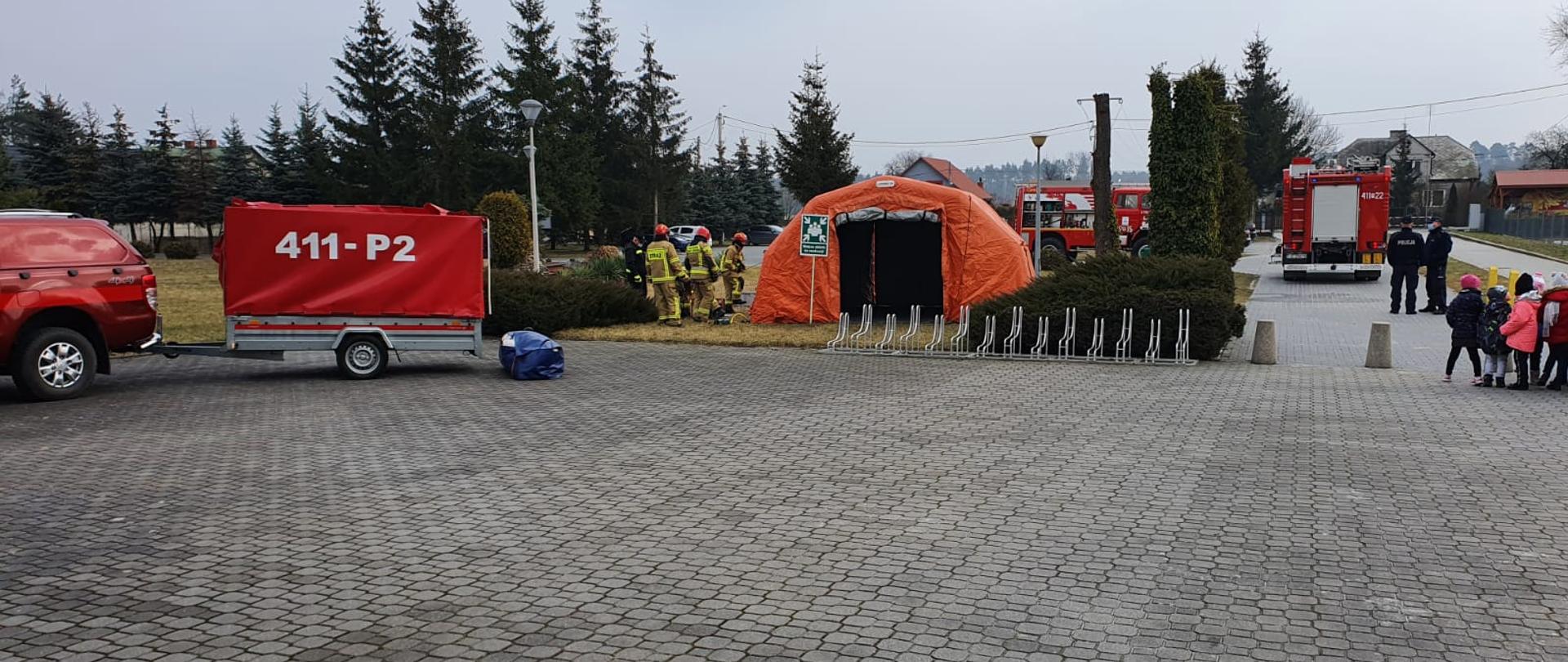 W centralnym punkcie zdjęcie widnieje namiot pneumatyczny w kolorze pomarańczowym. Przed nim znajduję się zielona tabliczka „Miejsca zbiórki do ewakuacji”. Tuż przy namiocie znajduje się kilku strażaków w ubraniach specjalnych, którzy pracują przy rozstawieniu namiotu. Po prawej stronie znajduję się kilkoro ewakuowanych dzieci, które podążają w kierunku namiotu. Po lewej stronie zaparkowany jest samochód lekki samochód rozpoznawczo-ratowniczy, do którego przypięta jest przyczepka w kolorze czerwonym. Na niej widnieje biały napis 411-P2. W tle zdjęcia widnieją dwa samochody ratowniczo-gaśnicze.