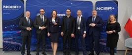 Przed niebieską ścianką z napisem NCBR stoi siedem osób, od lewej stoją wiceminister Gzik i minister Kulasek, w tle z prawej pod ścianą polska flaga.