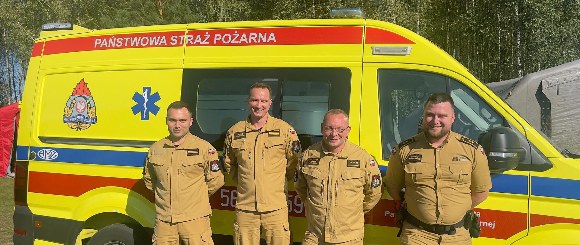 Przed żółtym ambulansem z napisem Państwowa Straż Pożarna stoi czterech mężczyzn ubranych w żółte mundury państwowej straży pożarnej