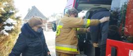 Fotografia przedstawiająca strażaka ochotnika pomagającego starszemu mężczyźnie wysiąść samochodu strażackiego z transportu do SPZOZ na szczepienia przeciw Covid-19 w ramach Narodowego Programu Szczepień. 